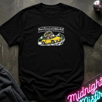 black graphic t-shirt laid flat on a dark textured surface. The graphic features a yellow sports car with a driver and passengers, with the text 'MAXIMUM EFFORT MINIMUM HORSEPOWER' above it.