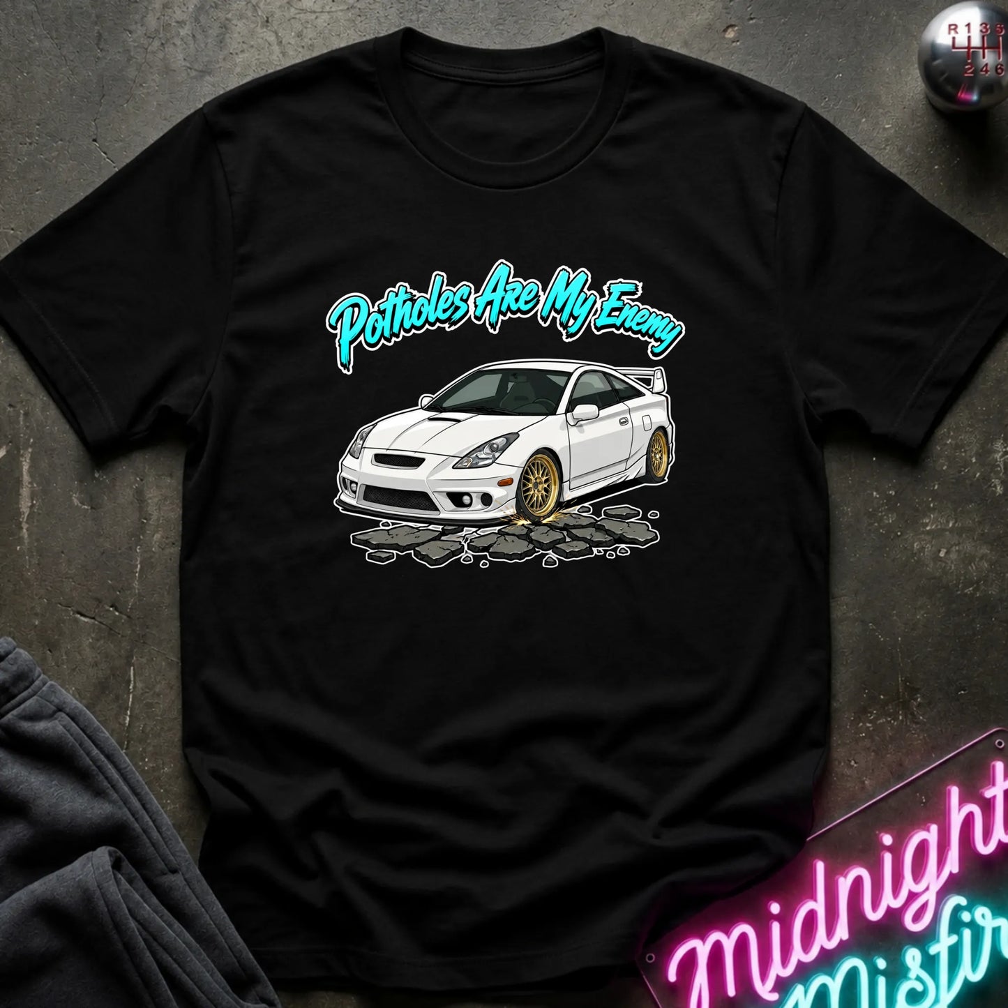 A black short-sleeve graphic t-shirt laid flat on a dark textured surface. The shirt features a white sports car with gold wheels and the text 'Potholes Are My Enemy' in a teal cursive font.