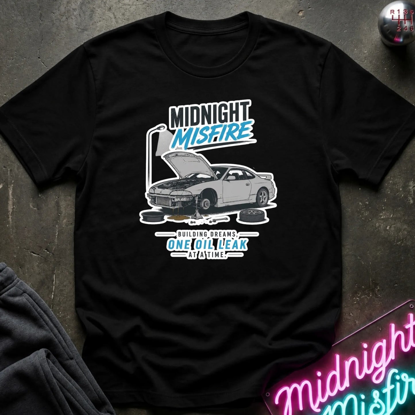 A black short-sleeve graphic T-shirt laid flat on a dark textured surface. The shirt features a white and blue graphic of a car with its hood open, surrounded by text reading 'MIDNIGHT MISFIRE' and smaller text below.