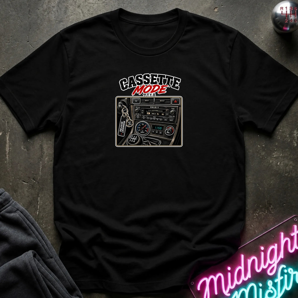 A black short-sleeve t-shirt laid flat on a dark textured surface, featuring a graphic print of a cassette radio with the text 'CASSETTE MODE' in black and red. A neon sign and dark pants are partially visible as props.