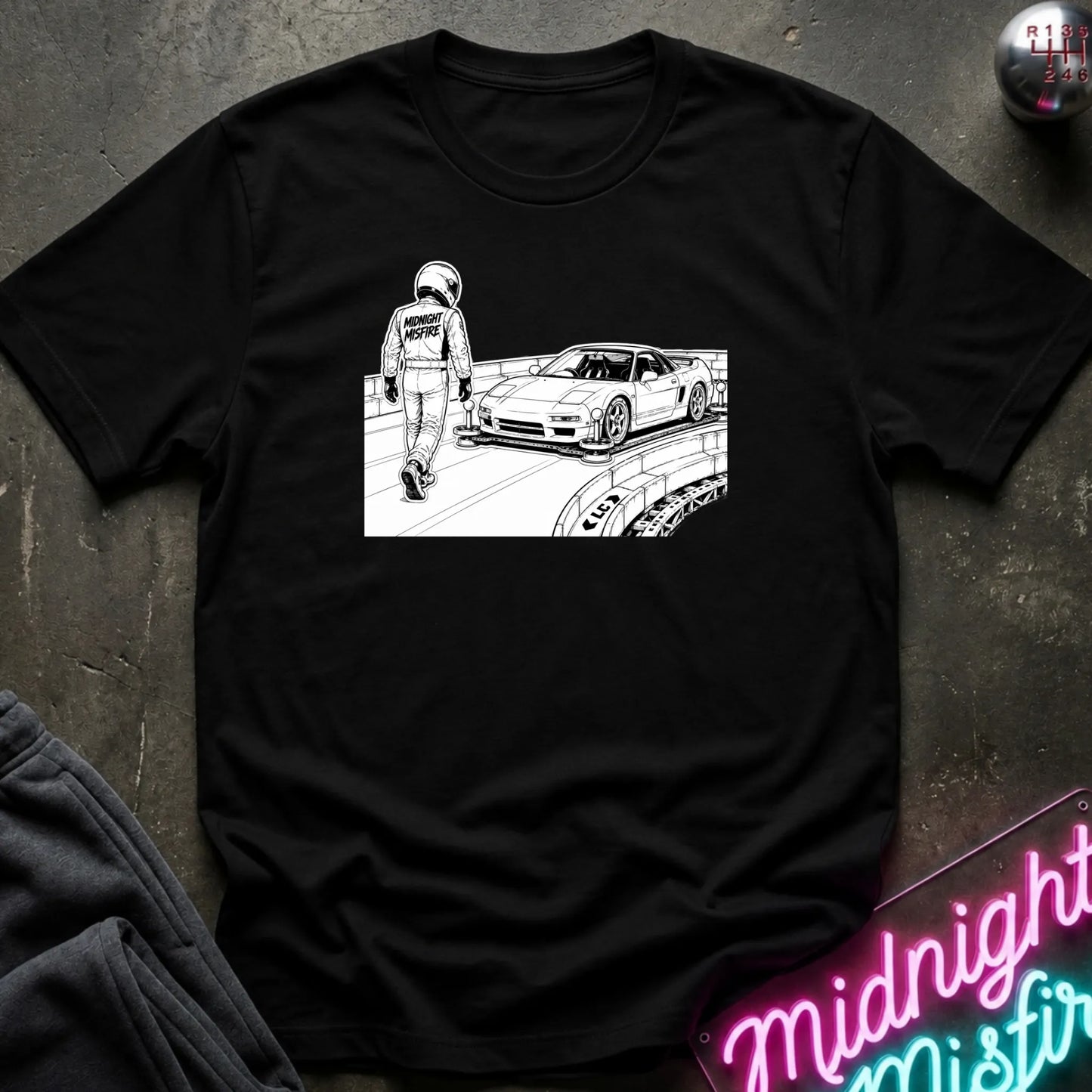 A black short-sleeve crewneck t-shirt laid flat on a dark textured surface. The shirt features a white line art graphic of a race car and a driver on a racetrack.