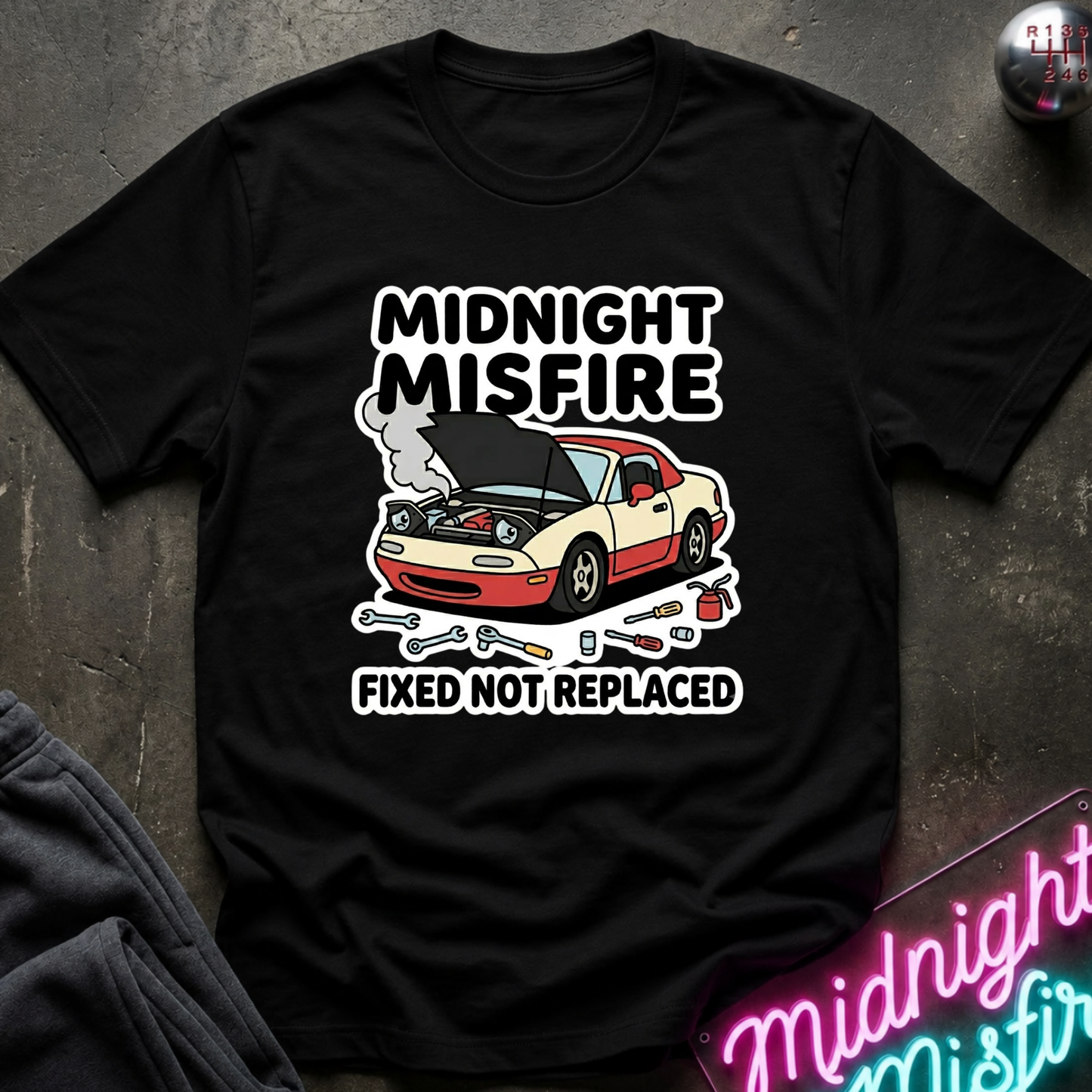 A black short-sleeve graphic T-shirt laid flat on a dark textured surface. The shirt features a cartoon car with its hood open, surrounded by tools, with the text 'MIDNIGHT MISFIRE' above and 'FIXED NOT REPLACED' below.