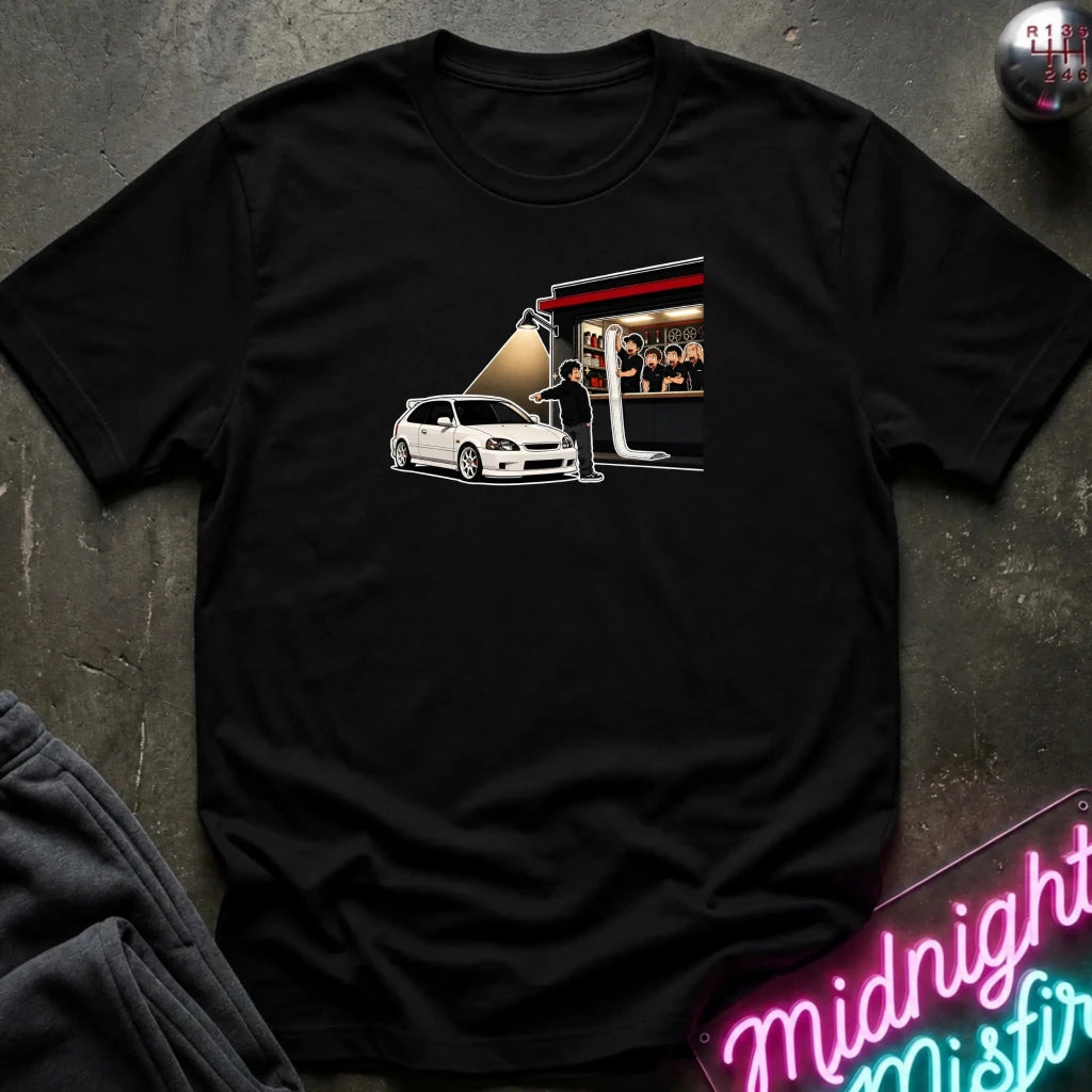A black short-sleeve graphic t-shirt laid flat on a dark textured surface. The graphic on the front shows a white hatchback car next to a stage-like scene with a spotlight 