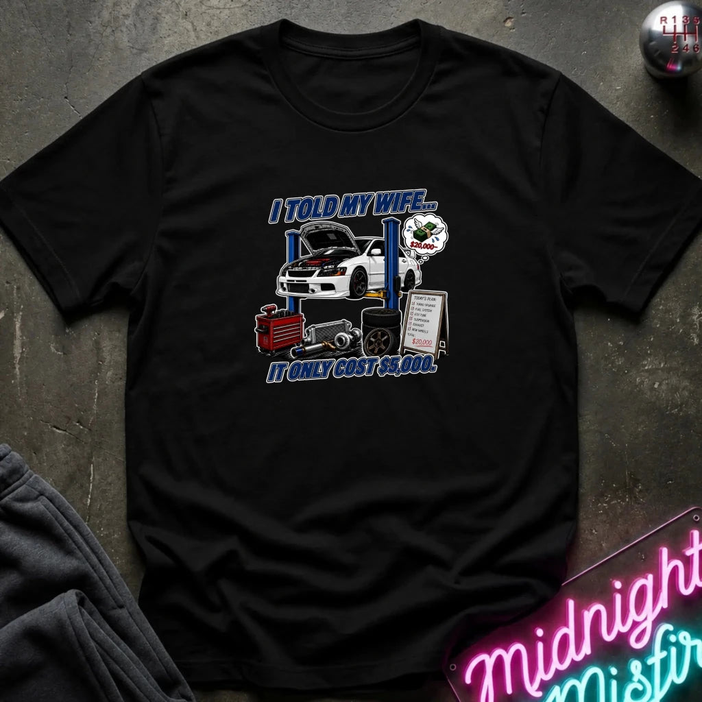 A black short-sleeve graphic t-shirt laid flat on a dark textured surface. The graphic features a white sports car on a blue lift with the text 'I TOLD MY WIFE.