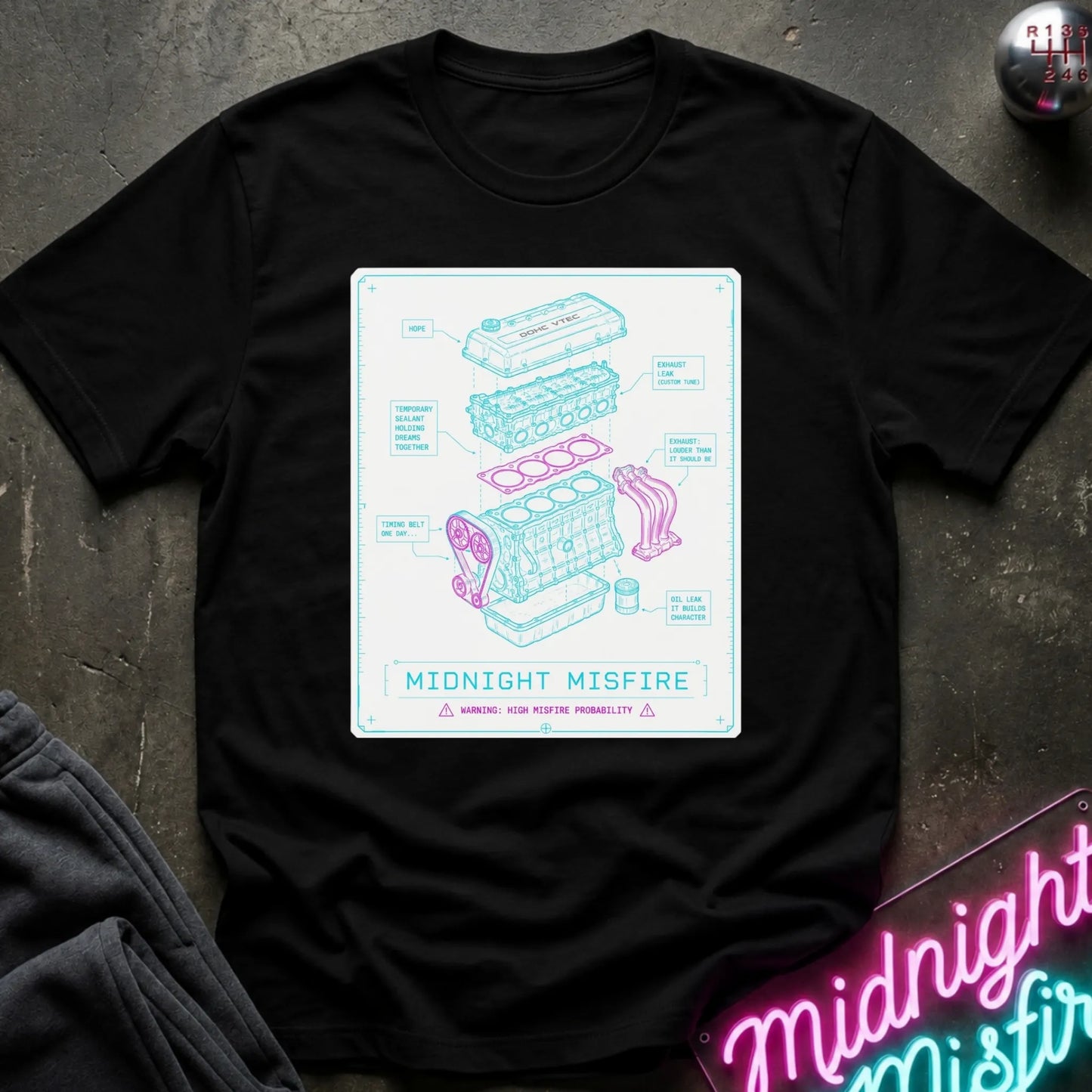 A black short-sleeve graphic T-shirt laid flat on a dark textured surface. The shirt features a large rectangular print with a light blue engine diagram and the text 'MIDNIGHT MISFIRE.