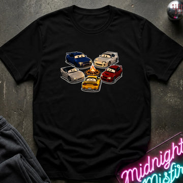A black short-sleeve t-shirt laid flat on a dark textured surface, featuring a graphic of cartoon race cars arranged around a campfire. The shirt is styled with a gray sweatshirt in the corner and a pink neon sign in the bottom right.