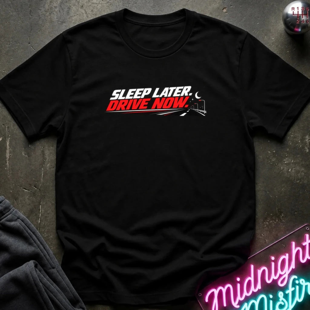 A black short-sleeve crewneck t-shirt laid flat on a dark textured surface. The shirt features a graphic print on the chest that reads 'SLEEP LATER.