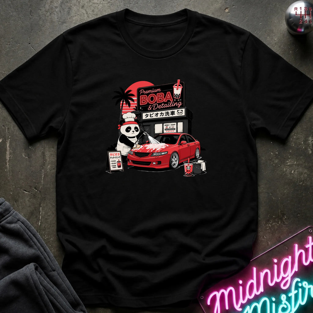 A black short-sleeve graphic T-shirt laid flat on a dark textured surface. The front print shows a red car, a panda character, and signage reading 'Premium DOBA Detailing' with Japanese text.
