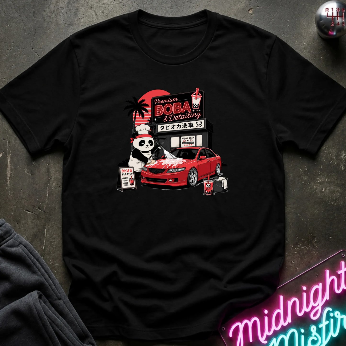 A black short-sleeve graphic T-shirt laid flat on a dark textured surface. The front print shows a red car, a panda character, and signage reading 'Premium DOBA Detailing' with Japanese text.
