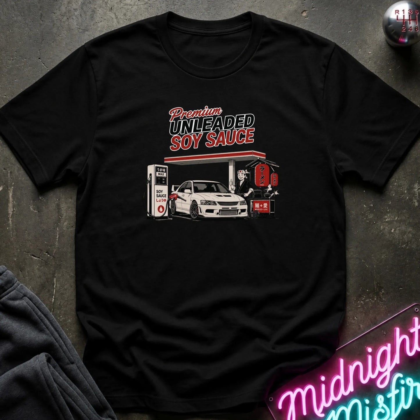 A black short-sleeve graphic t-shirt laid flat on a dark textured surface. The graphic features a white car at a gas station with the text 'Premium UNLEADED SOY SAUCE' in red and white.
