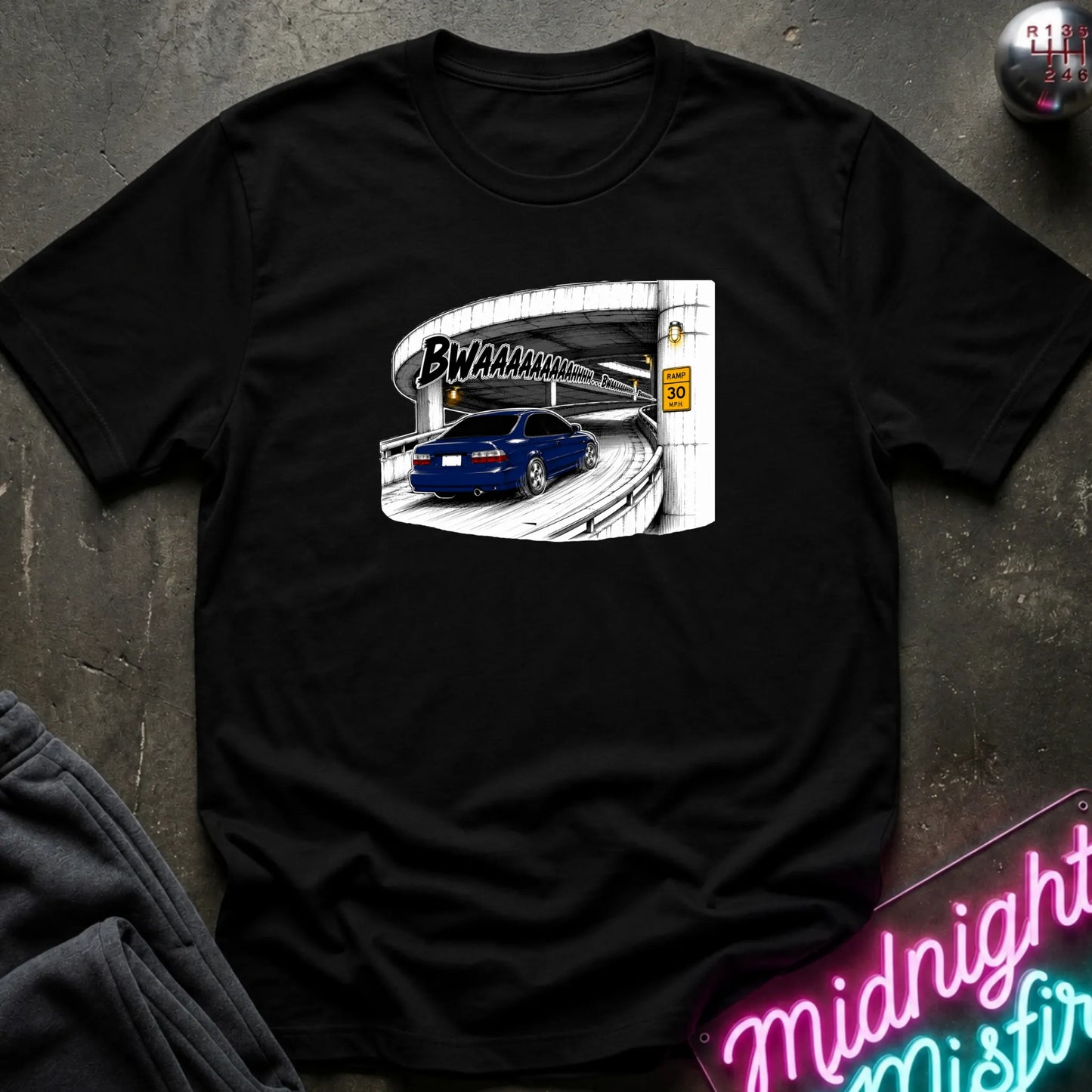 A black short-sleeve crewneck t-shirt laid flat on a dark textured surface. The shirt features a graphic print of a blue car driving under a highway overpass with the text 'BWAHAHA'.