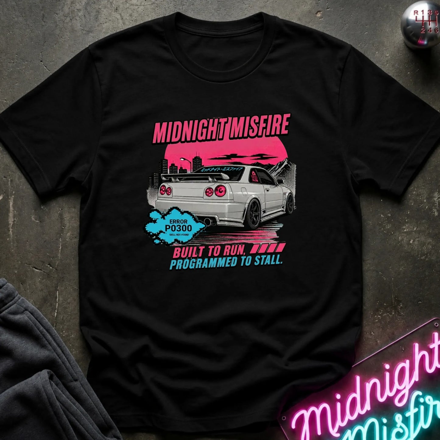 black short-sleeve graphic t-shirt laid flat on a dark textured surface. The graphic features a white sports car with a pink sunset, the text 'MIDNIGHT MISFIRE' in pink.
