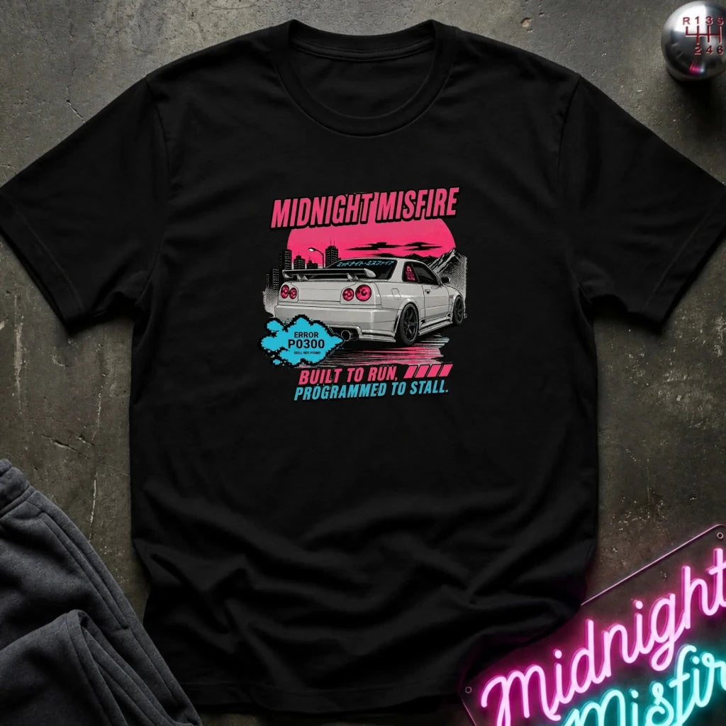 A black short-sleeve graphic t-shirt laid flat on a dark textured surface. The graphic features a silver car against a pink background with the text 'MIDNIGHTMISFIRE' 