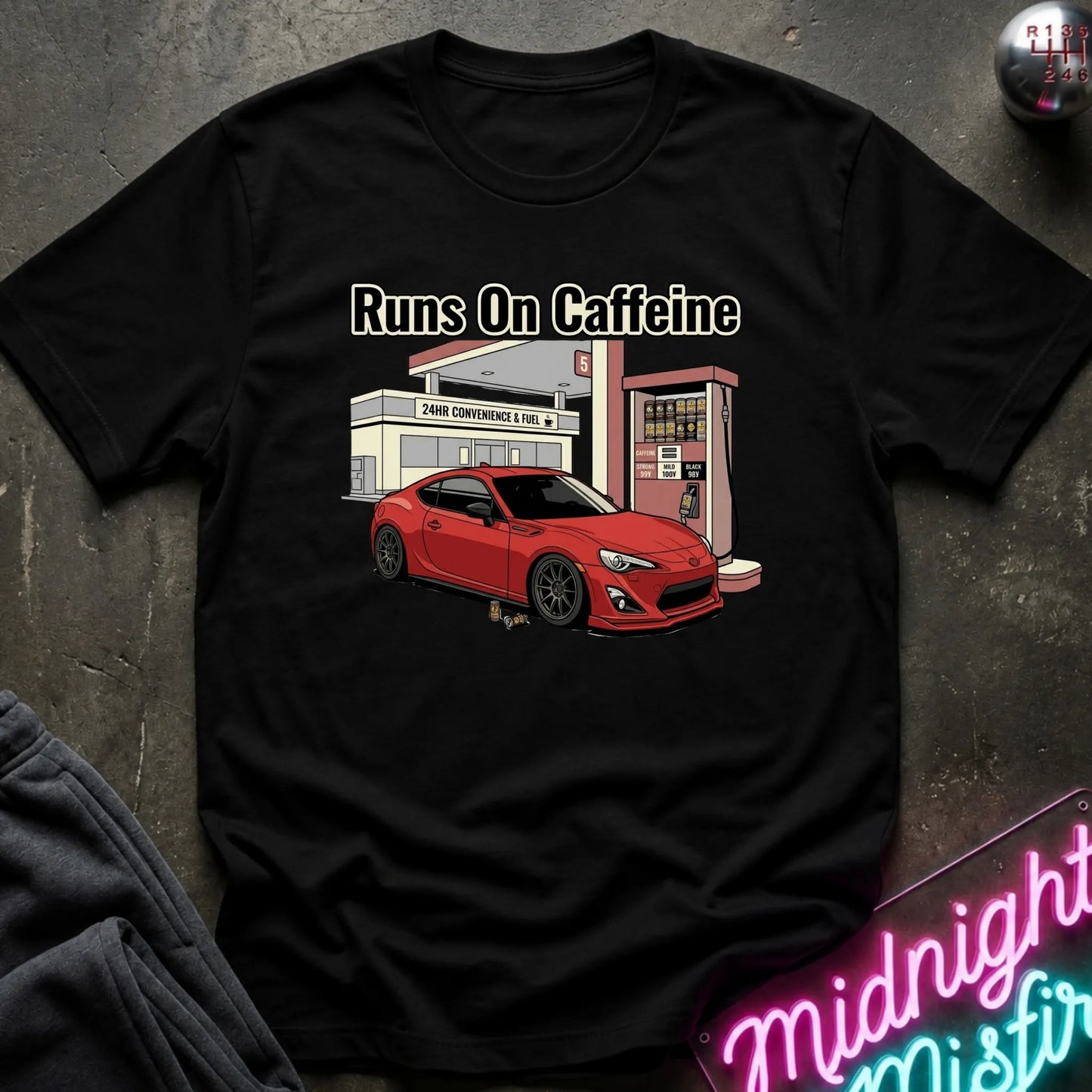 black short-sleeve graphic T-shirt laid flat on a dark textured surface. The shirt features a printed illustration of a red sports car parked in front of a convenience store and gas station, with the text 'Runs On Caffeine' above the graphic.