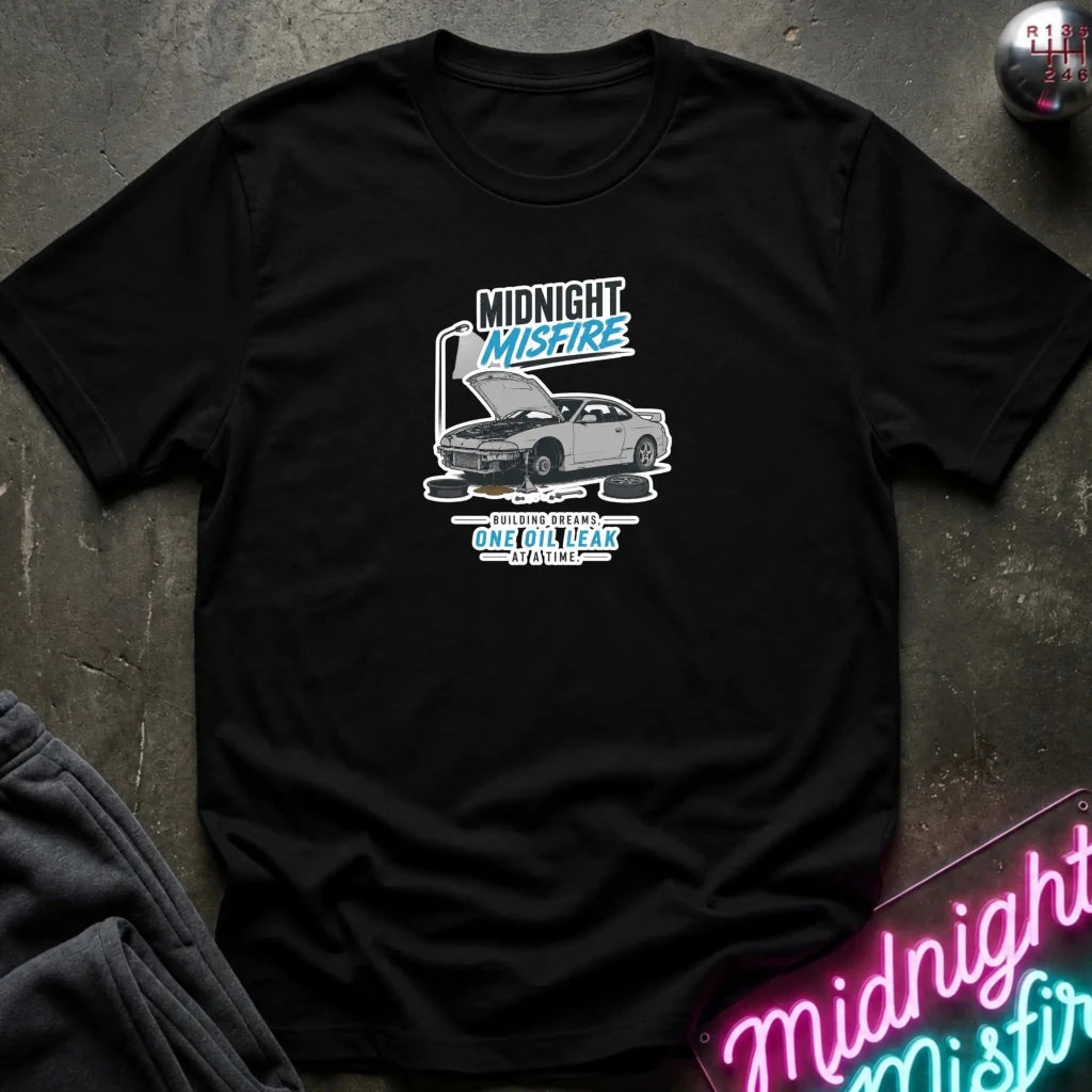 A black short-sleeve graphic t-shirt laid flat on a dark textured surface. The graphic features a silver sports car with its hood open, accompanied by the text 'MIDNIGHT MISFIRE'
