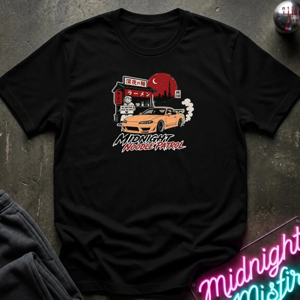 black crewneck t-shirt featuring a graphic print of a yellow Nissan Silvia S15 drift car in front of a red moon and Japanese storefront, with the text 'MIDNIGHT Noodle PATROL'. The shirt is laid flat on a dark textured surface with a neon sign and dark pants partially visible.
