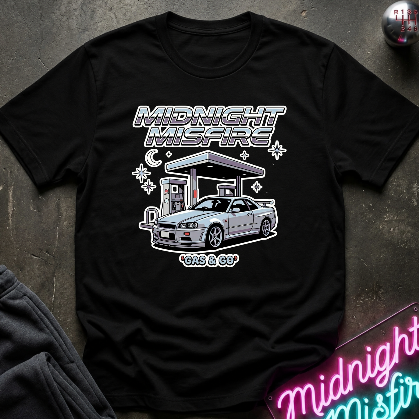 A black short-sleeve graphic t-shirt laid flat on a dark textured surface. The shirt features a white and gray graphic print of a sports car at a gas station with the text 'MIDNIGHT MISFIRE' and 'GAS GO'.
