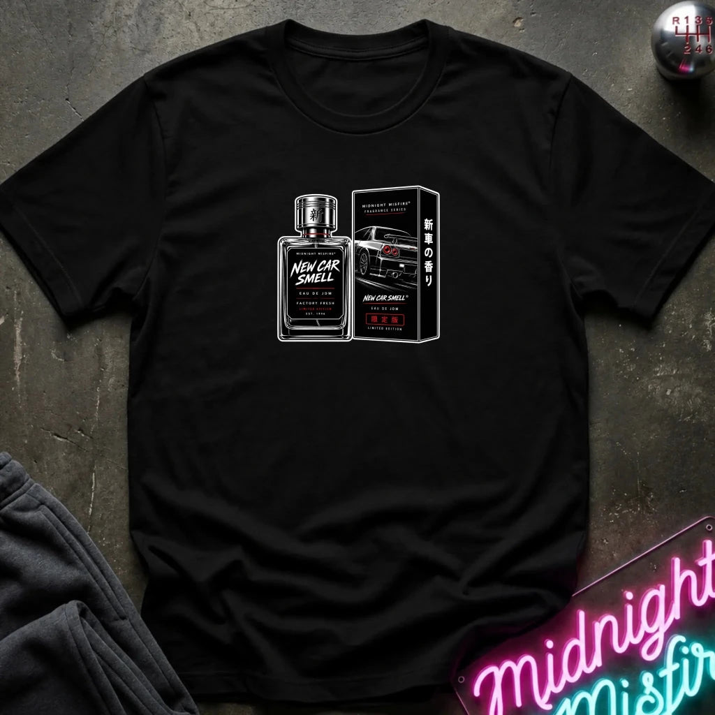 A black short-sleeve graphic t-shirt laid flat on a dark textured surface. The shirt features a white and red graphic print of a perfume bottle and its box labeled 'New Car Smell'.