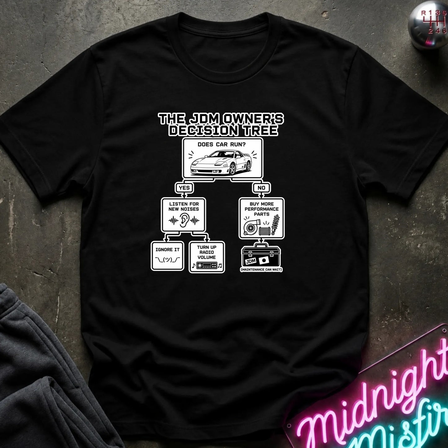 A black short-sleeve graphic T-shirt laid flat on a dark textured surface. The shirt features a black printed decision tree titled 'THE JDM OWNER'S DECISION TREE' with illustrations and text related to car care and maintenance.