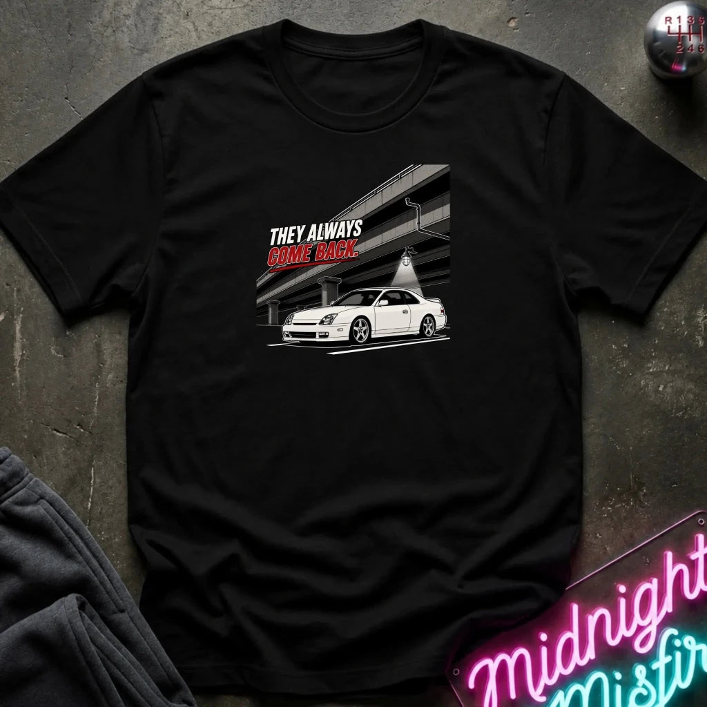 A black short-sleeve graphic t-shirt laid flat on a dark textured surface. The graphic features a white sports car under an elevated highway with the text 'THEY ALWAYS COME BACK.