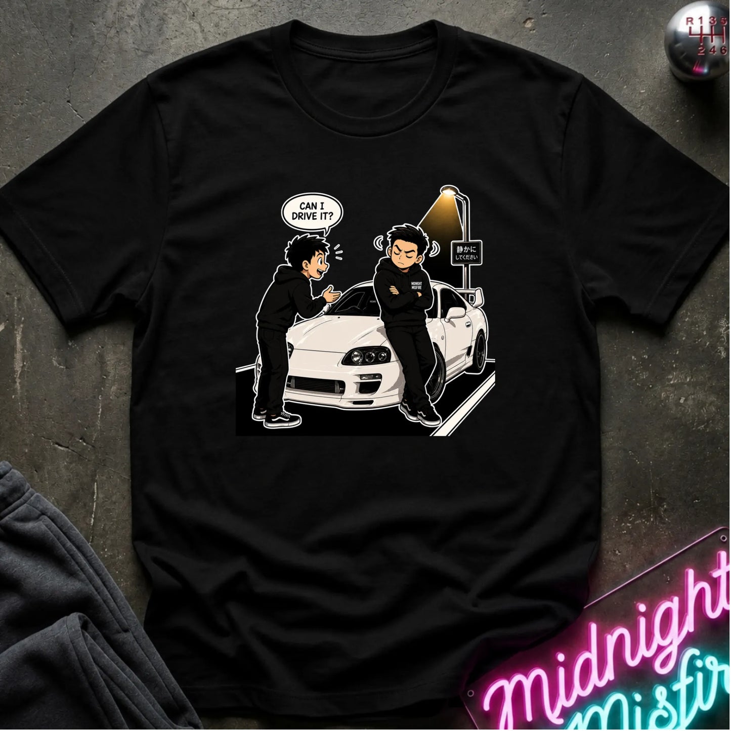 A black short-sleeve graphic t-shirt laid flat on a dark textured surface. The graphic shows a white sports car with two illustrated figures standing beside it, one with a speech bubble reading 'CAN I DRIVE IT?'.