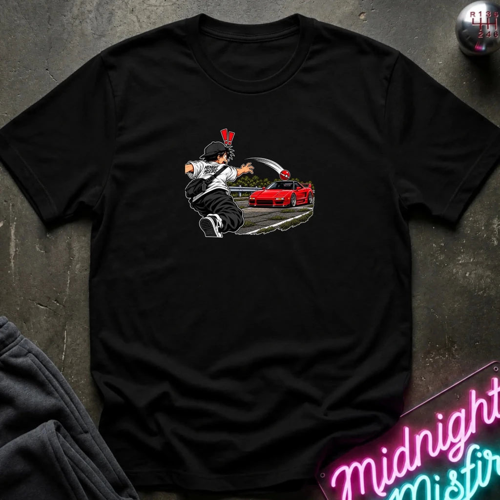 A black short-sleeve crewneck t-shirt laid flat on a dark textured surface. The center of the shirt features a graphic print of an anime character in a black and white outfit with a hat, next to a sports car 