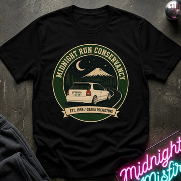A black short-sleeve graphic T-shirt laid flat on a dark textured surface. The shirt features a circular vintage-style print with the words 'MIDNIGHT RUN CONSERVANCY,' a white car on a winding road, a mountain under a crescent moon, and a banner reading 'EST.