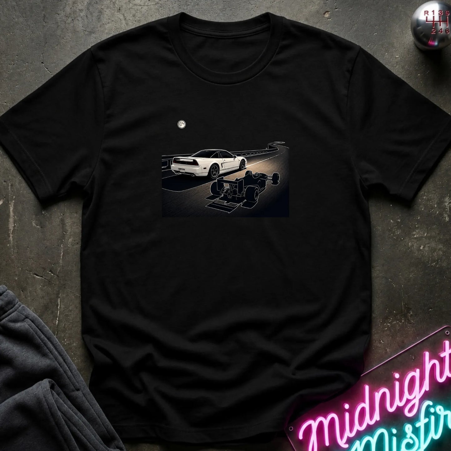 A black short-sleeve crewneck t-shirt laid flat on a dark textured surface, featuring a graphic print of a white sports car and a shadow car on a night road scene. The shirt is styled with dark gray sweatpants, a silver ball, and a neon sign in the corners.