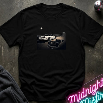 A black short-sleeve crewneck t-shirt laid flat on a dark textured surface, featuring a graphic print of a white sports car and a shadow car on a night road scene. The shirt is styled with dark gray sweatpants, a silver ball, and a neon sign in the corners.