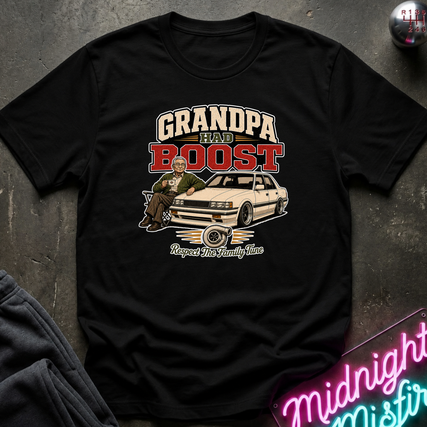 A black short-sleeve graphic t-shirt laid flat on a dark textured surface. The graphic features the text 'GRANDPA HAD BOOST' in cream and red lettering above an illustration of a man sitting in a white car with the phrase 'Respect The Family Line' below.