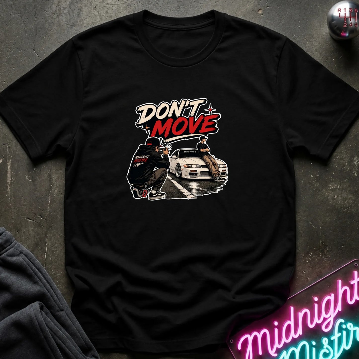 A black short-sleeve graphic t-shirt laid flat on a dark textured surface. The graphic features the text 'DON'T MOVE' in stylized white and red lettering above an illustration of a person crouching next to a white sports car.