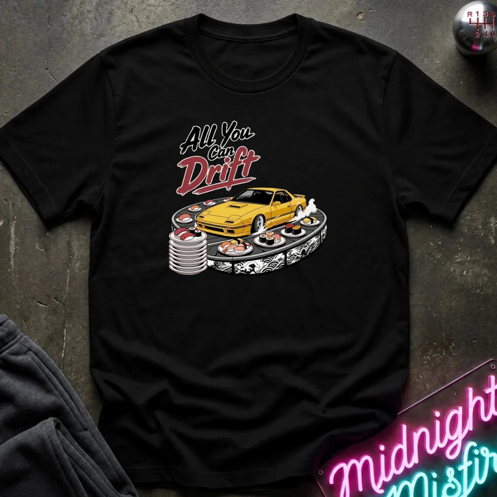 A black short-sleeve graphic t-shirt laid flat on a dark textured surface. The graphic features a yellow sports car surrounded by stacks of sushi and the text 'All You Can Drift' in stylized white and red lettering.