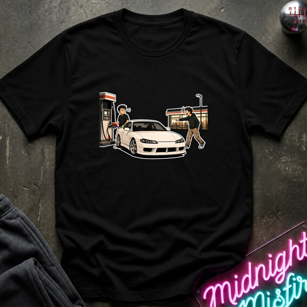 A black short-sleeve graphic t-shirt laid flat on a dark textured surface. The graphic shows a white sports car at a gas station with two illustrated figures.