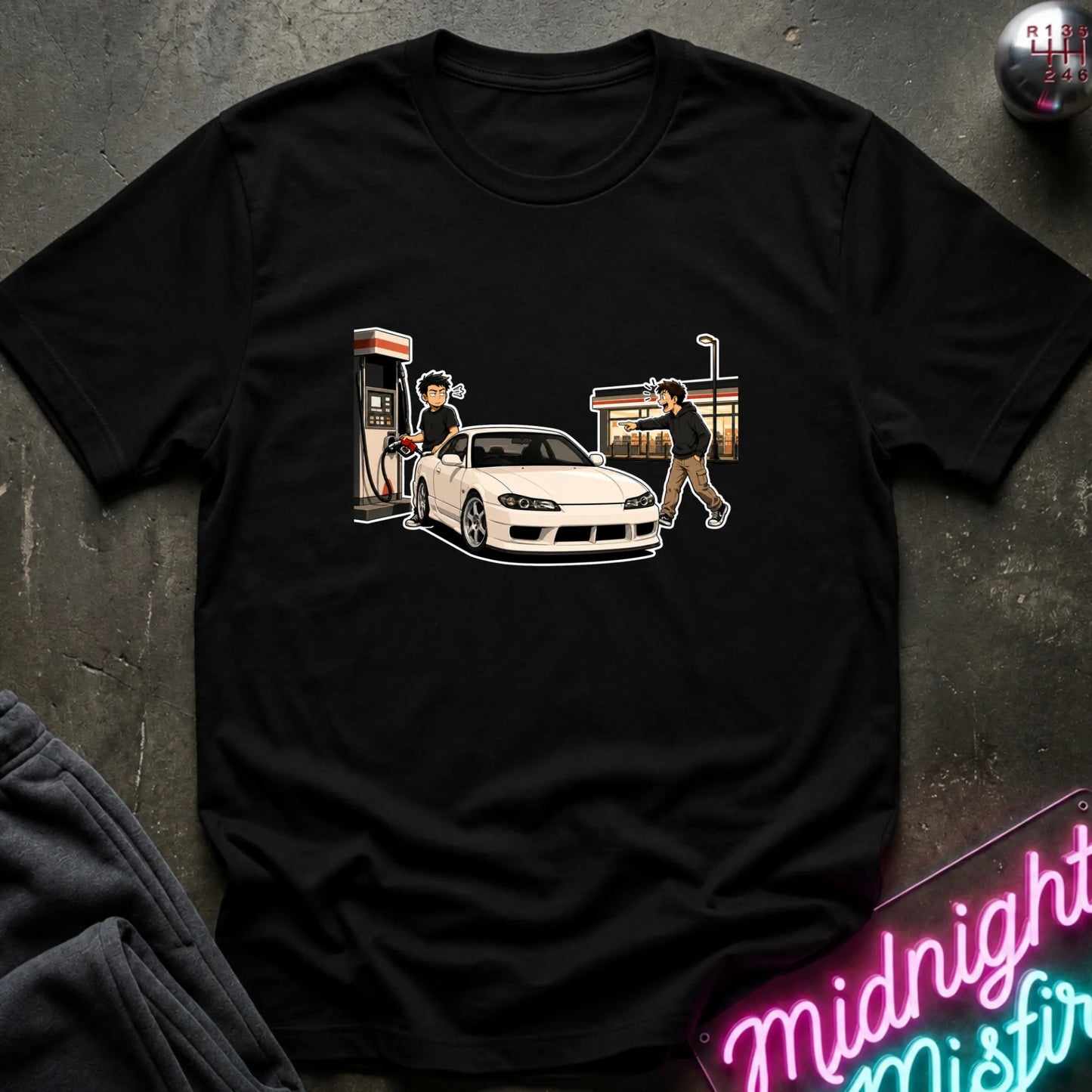 A black short-sleeve graphic t-shirt laid flat on a dark textured surface. The graphic shows a white sports car at a gas station with two illustrated figures.