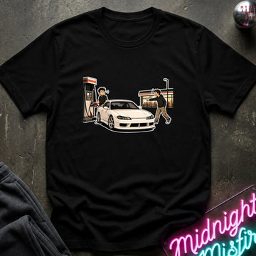A black short-sleeve graphic t-shirt laid flat on a dark textured surface. The graphic shows a white sports car at a gas station with two illustrated figures.