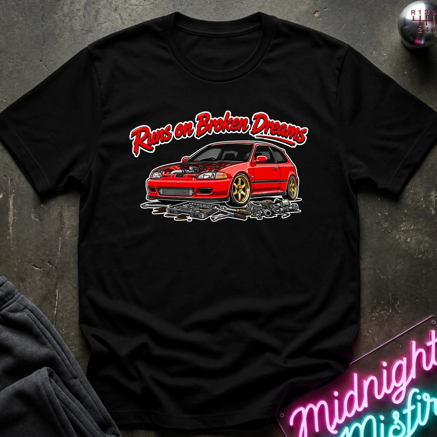 black graphic t-shirt laid flat on a dark textured surface. The shirt features a red sports car with gold wheels and the text 'Runs on Broken Dreams' in red and white.