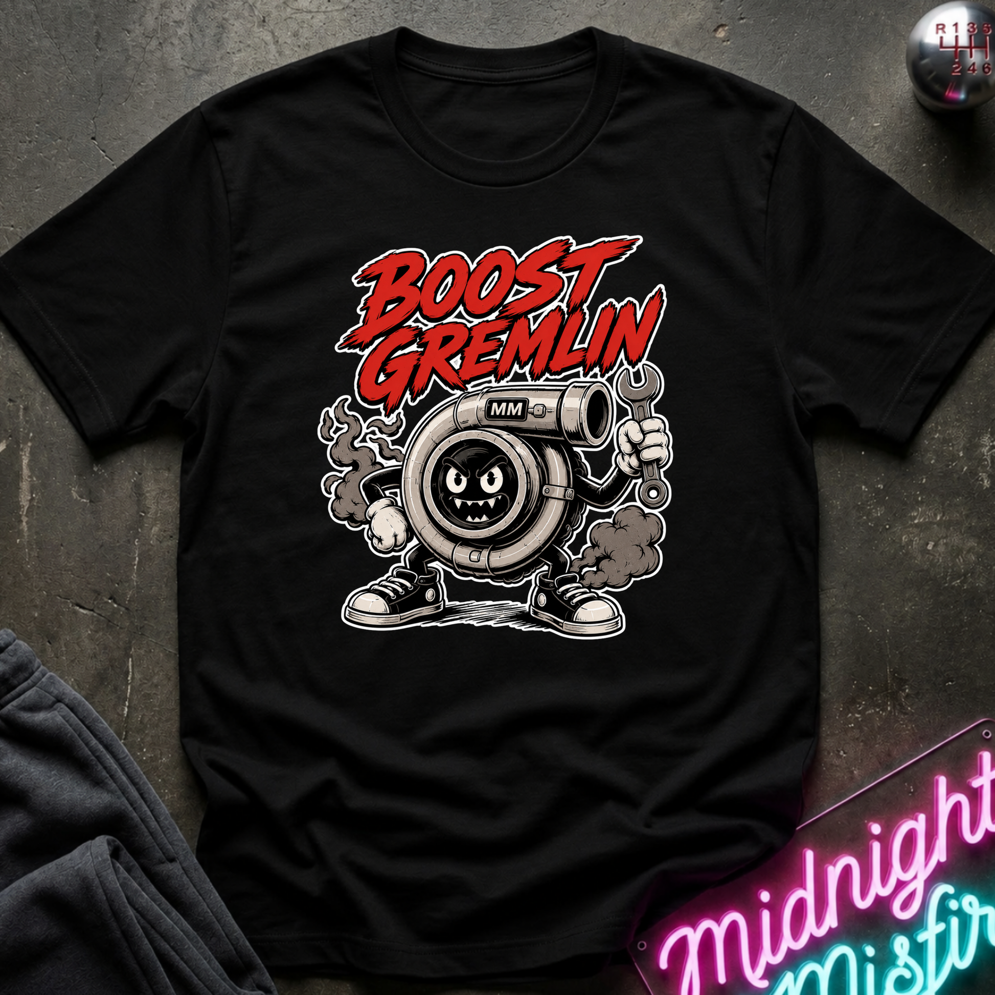 A black short-sleeve graphic T-shirt laid flat on a dark textured surface. The shirt features a large front print with the words 'BOOST GREMLIN' in red and white, above a cartoon turbocharger character in gray, white, and black.