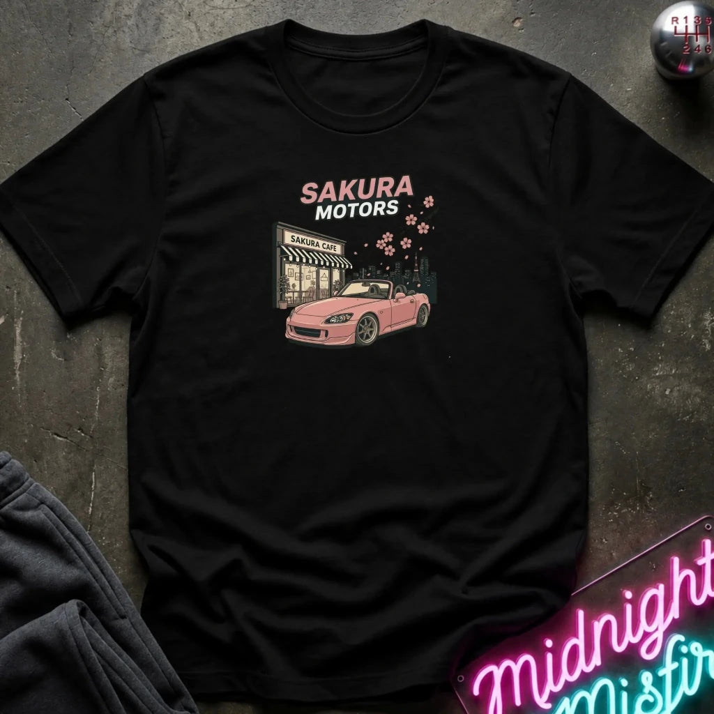 A black short-sleeve graphic t-shirt laid flat on a dark textured surface. The shirt features a pink convertible car and a storefront with the text 'SAKURA MOTORS' in pink and white, accented by small pink cherry blossoms.