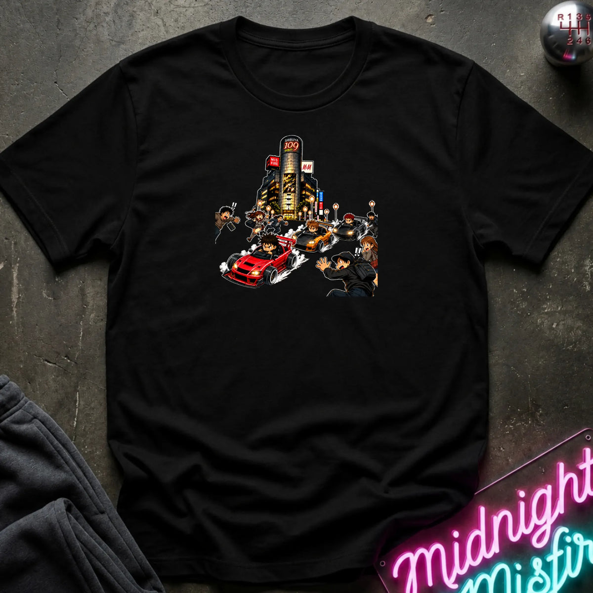 A black short-sleeve crewneck t-shirt with a graphic print of a city street scene featuring a red sports car and small figures, laid flat on a dark textured surface with a neon sign and dark pants partially visible.