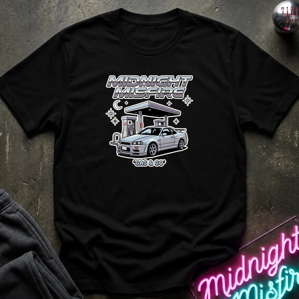 A black short-sleeve t-shirt with a graphic print of a silver Nissan S13 'Kits & Co' car at a gas station under the text 'MIDNIGHT MISFIRE' and 'gas & GO', laid flat on a dark textured surface.
