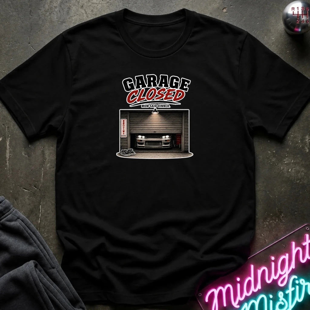 A black  short-sleeve graphic t-shirt laid flat on a dark textured surface. The graphic features the text 'GARAGE CLOSED' in black and red, above an illustration of a car parked inside a garage.