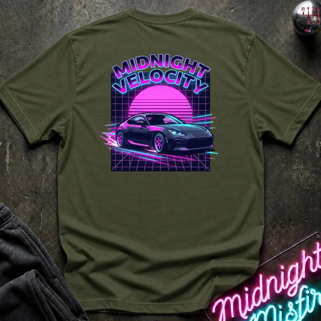 An olive green short-sleeve t-shirt laid flat on a dark textured surface, featuring a colorful retro 1980s-style graphic on the back with the text 'MIDNIGHT VELOCITY', a pink sun, and a dark sports car.