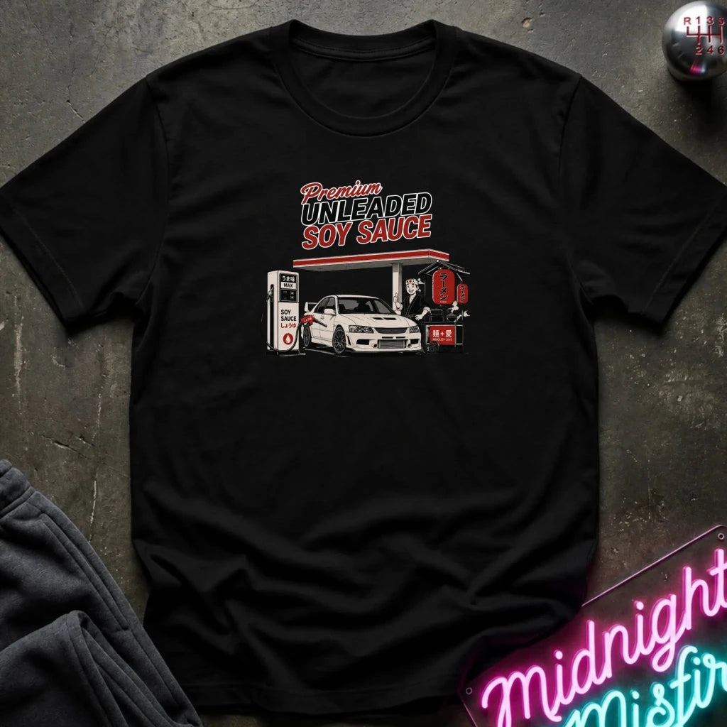 A black short-sleeve graphic t-shirt laid flat on a dark textured surface. The graphic features the text 'Premium UNLEADED SOY SAUCE' above an illustration of a white sports car at a gas station with a red sign reading '95'.