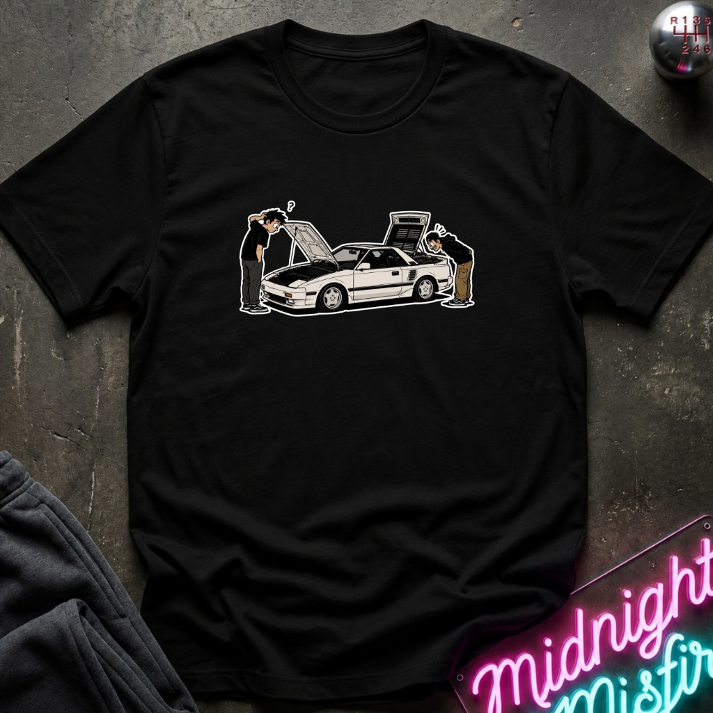 A black short-sleeve t-shirt laid flat on a dark textured surface, featuring a graphic print of a white sports car with two people inspecting it. A neon sign reading 'Midnight Mist' is partially visible in the bottom right corner.