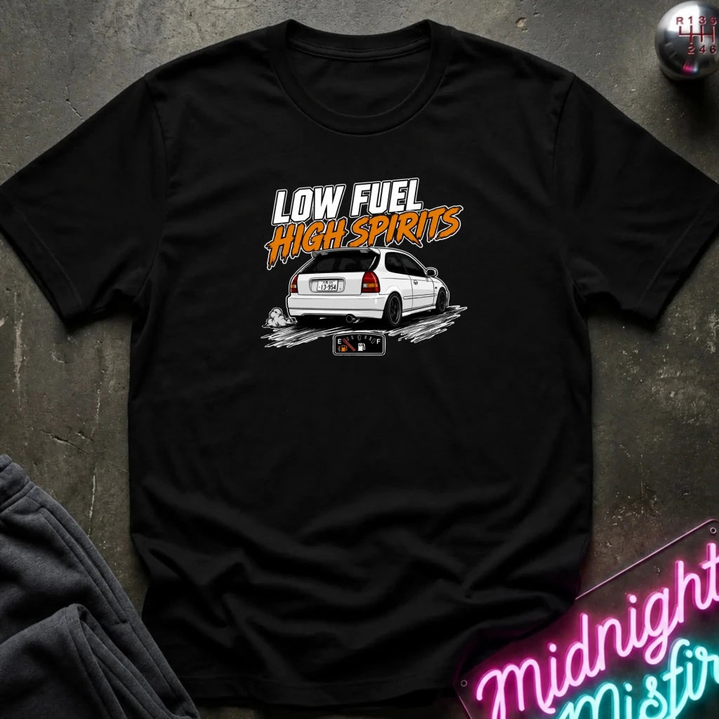 A black short-sleeve graphic t-shirt laid flat on a dark textured surface. The graphic features the text 'LOW FUEL HIGH SPIRITS' in white and orange above an illustration of a white hatchback car with smoke effects.