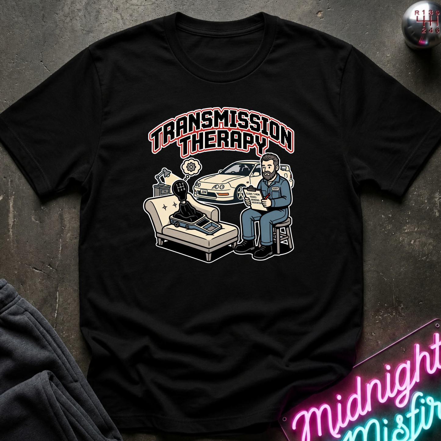 A black short-sleeve graphic t-shirt laid flat on a dark textured surface. The shirt features a 'Transmission Therapy' cartoon illustration with a therapist, a car, and a transmission.