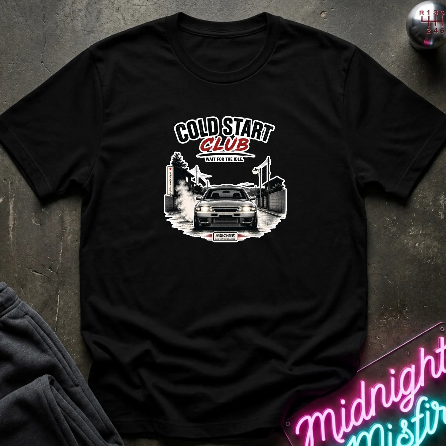 A black short-sleeve graphic t-shirt laid flat on a dark textured surface. The shirt features a white and red graphic print with the text 'COLD START CLUB' and an illustration of a car.