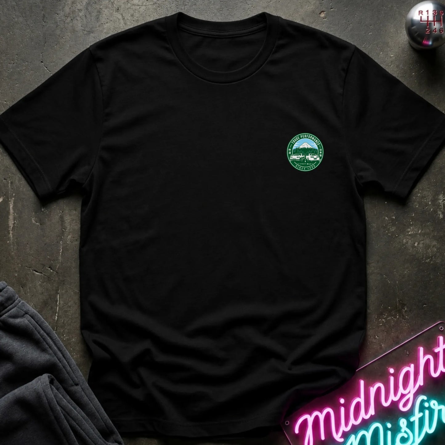A black short-sleeve graphic T-shirt laid flat on a dark textured surface. The shirt features a small circular green and white print logo  with a mountain, two cars, and the text 'TOUGE PERFORMANCE' and 'SINCE 1990'.
