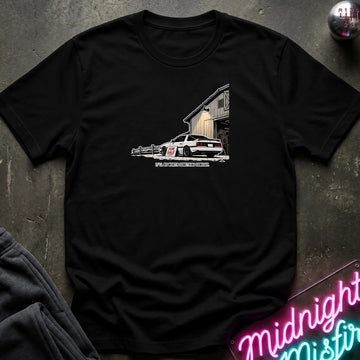 A black short-sleeve graphic t-shirt laid flat on a dark textured surface. The graphic shows a white sports car in front of a barn 