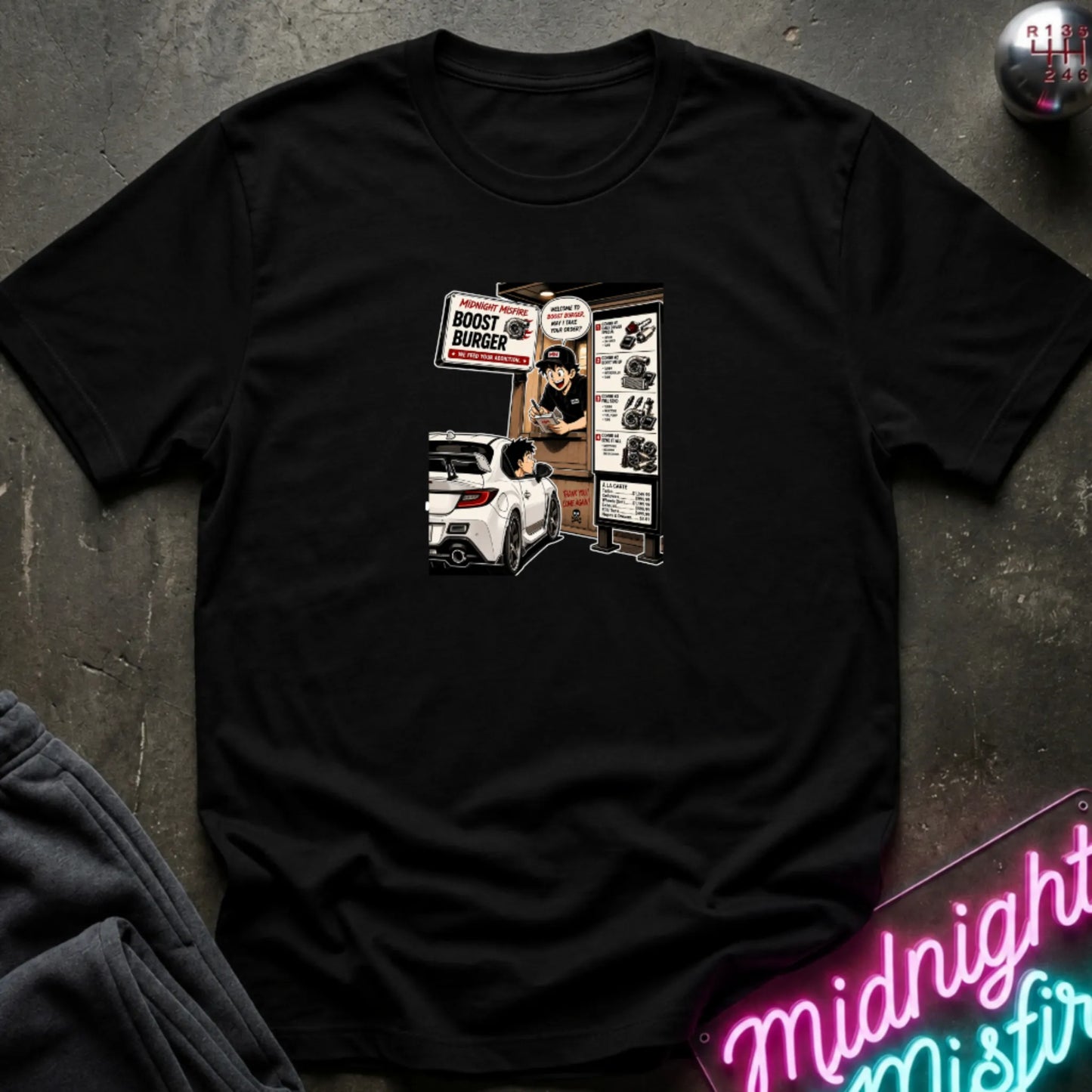 A black  short-sleeve crewneck t-shirt laid flat on a dark textured surface, featuring a central graphic print of a street-food burger scene with a white car and signs. 